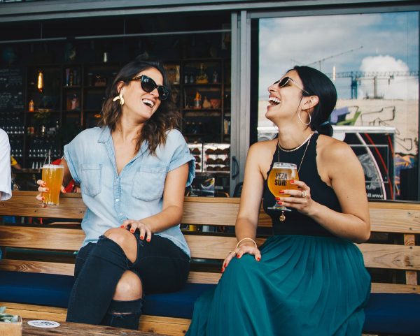 2 Girls Laughing While Holding a Beer and Sitting outside a Bar