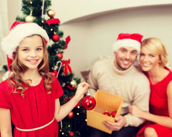 Dad holding a box with red Bauble and Mom holding the husband while daughter is standing infront of them holding a single red bauble and wearing a santa hat