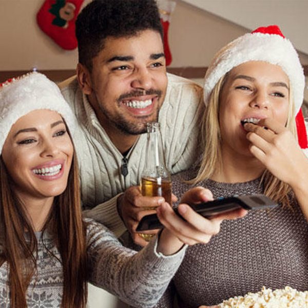 Three friends enjoying watching christmas movies with the other man holding a beer and both girls wearing santa hat