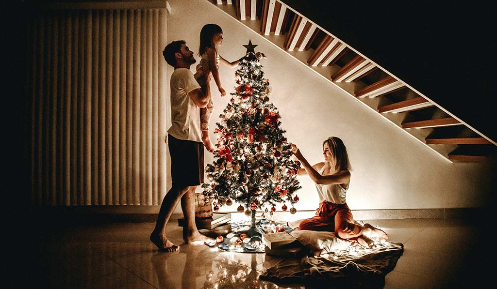 Father lifting his Daughter to add a star in a Christmas tree and wife adding some decor to the tree as well