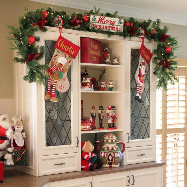 A Christmas Kitchen Hutch cabinet filled with Christmas Ornaments and decor also a hanging gingerbread stockings and Garlands