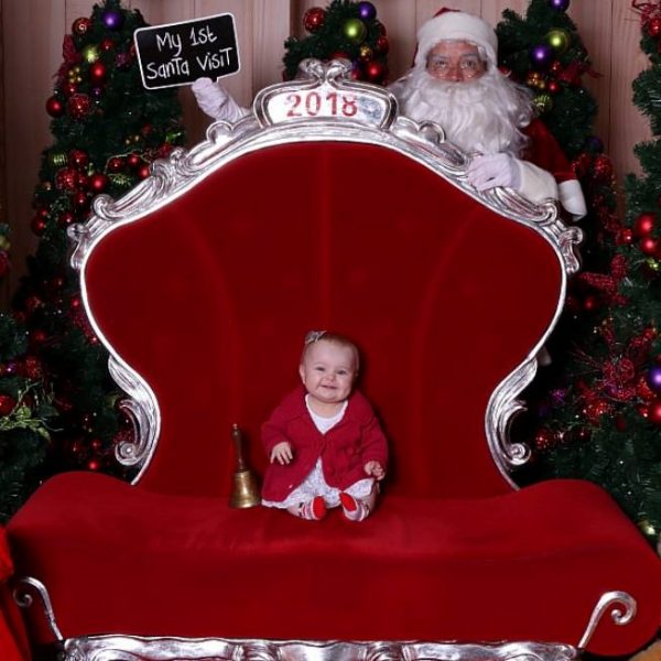 Cute litte girl sitting in Santas Big Red Chair While Santa Is behind holding a sign saying my 1st Santa Visit