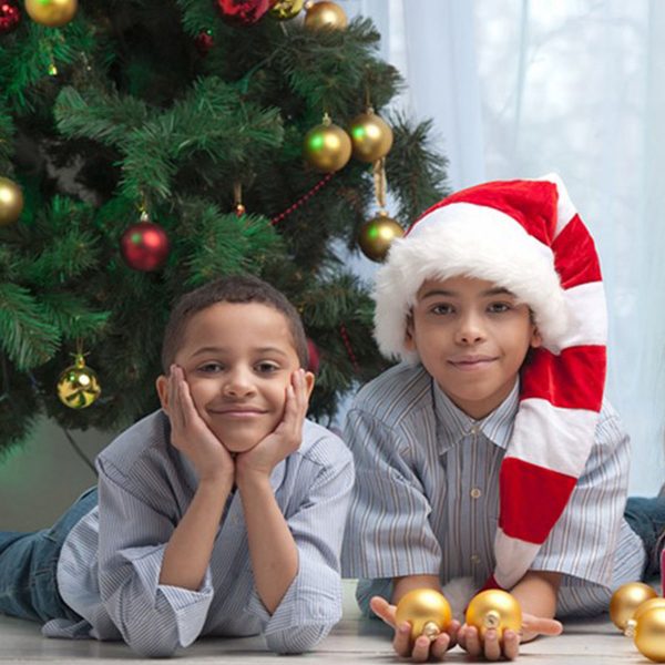 2 kids sitting in front of the Christmas Tree one kid is wearing a santa hat while holding a golden Bauble