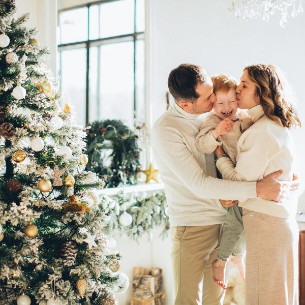 A man and a woman hugging their son and giving him a kiss wearing sweater, they are standing beside a big Christmas Tree