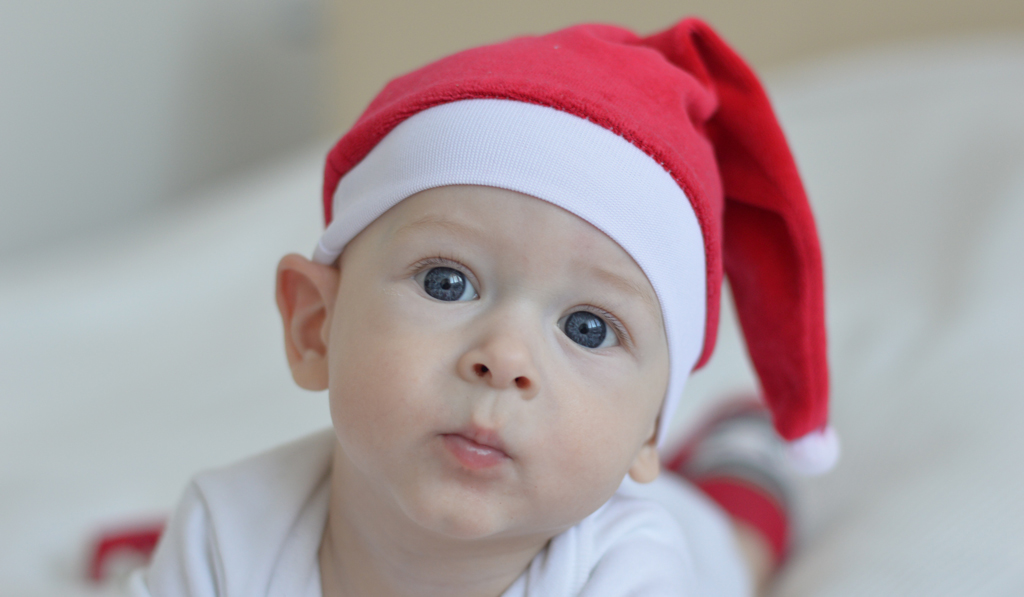 A baby crawling staring at the camera with blue eyes and wearing a red Santa hat
