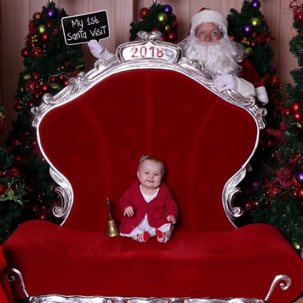 Baby girl wearing her red sweater sitting in the red chair while santa claus is holding the sign My Ist Santa visit